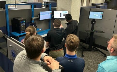 A group of people in an office environment gathered around a set of computer monitors and equipment, receiving training on a Manufacturing Execution System (MES).