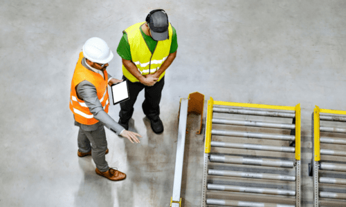 Two workers in a manufacturing facility, wearing safety vests and helmets, discussing operations near a conveyor system. One worker is holding a tablet, demonstrating the use of real-time data for decision-making.